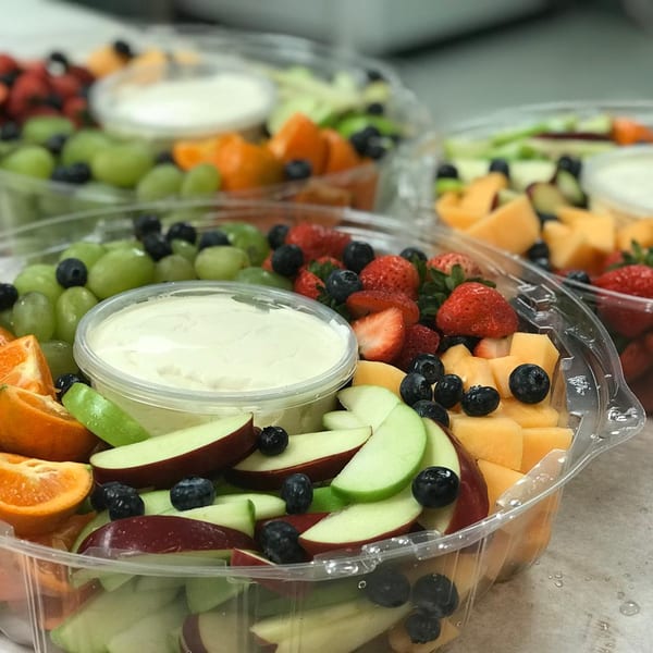 Several clear plastic containers of fresh fruit, including grapes, strawberries, blueberries, apples, and melon.