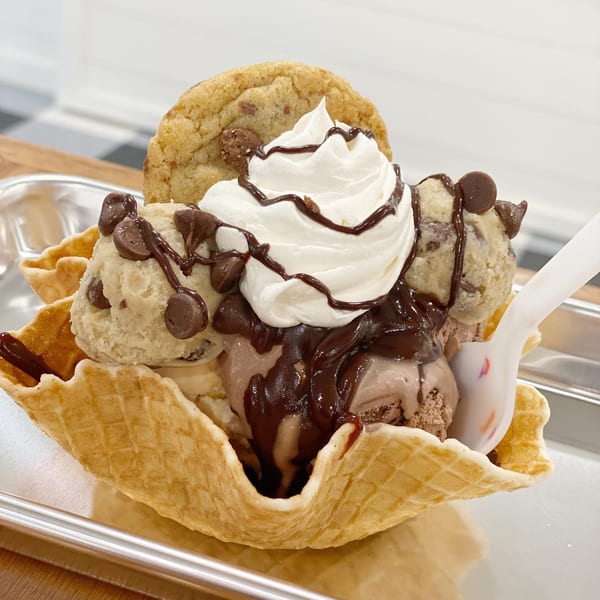 A waffle bowl sundae with cookie dough and chocolate ice cream, whipped cream, chocolate drizzle, and a cookie.