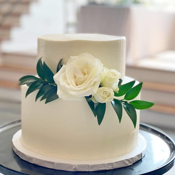 Two-tiered white wedding cake adorned with white roses and green leaves on a silver tray.