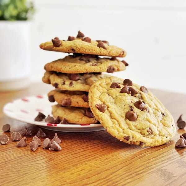 A stack of five chocolate chip cookies on a white plate, with loose chips on a wooden table.